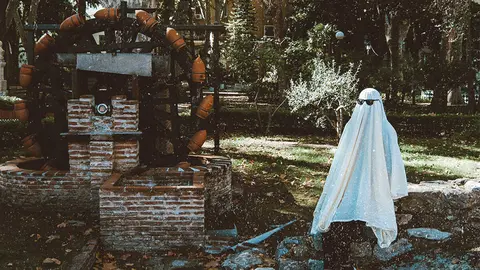 Fantasma moderno con gafas de sol posando junto a antigua noria de agua de ladrillo en un parque de Algeciras, ambiente oto&ntilde;al y misterioso.